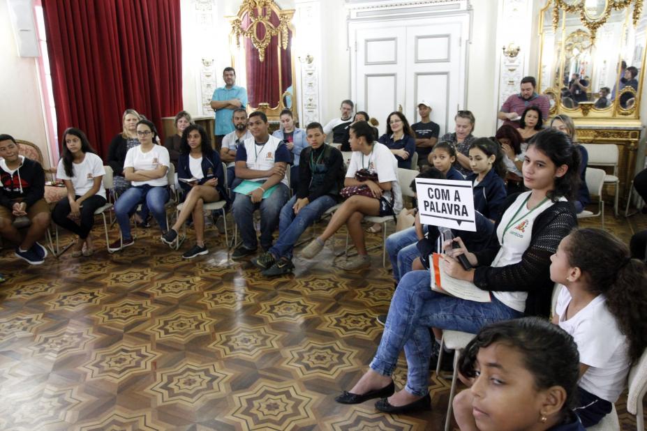 Alunos em círculo durante mediação. Uma delas segura placa dizendo com a palavra #pracegover 