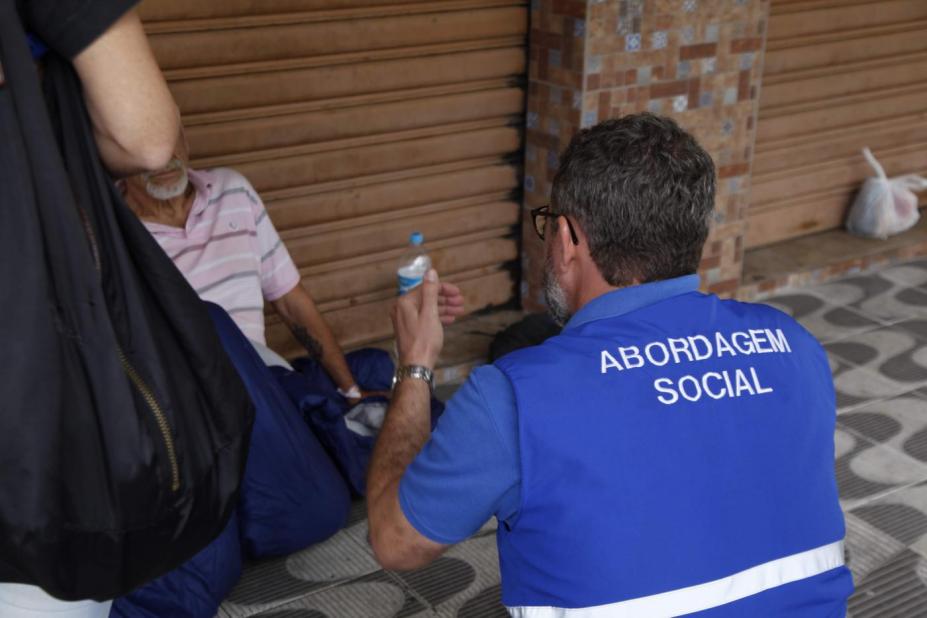 agente usando colete onde se lê nas costas abordagem social, conversa com homem que está sentado no chão de uma rua. #paratodosverem