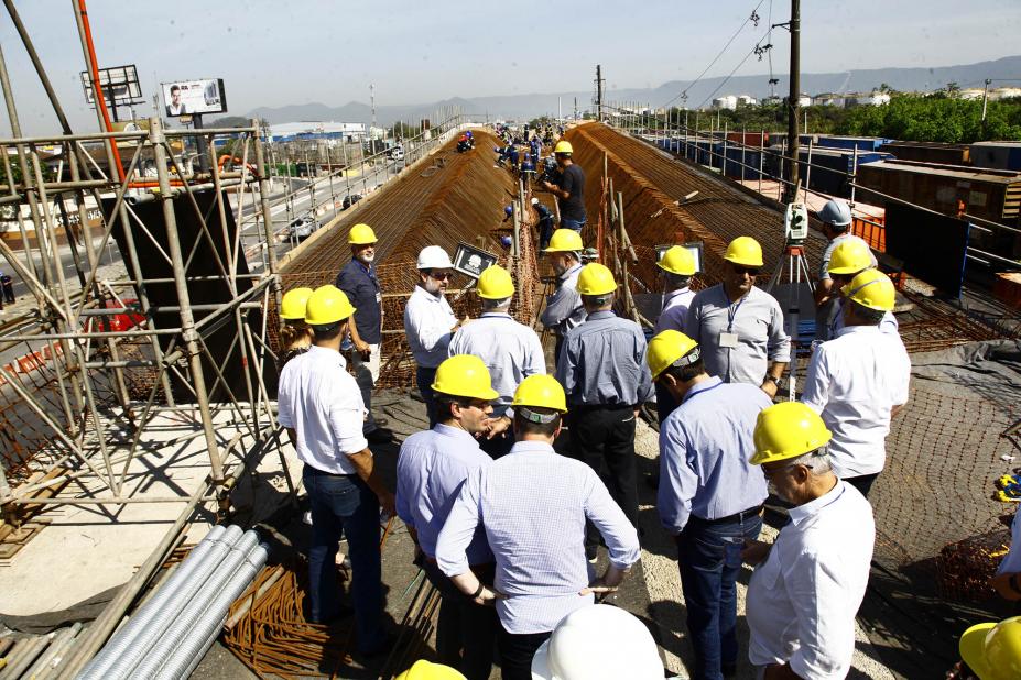 Homens e mulheres usando capacetes estão diante da obra de uma ponte. Há uma grande estrutura de ferros. #Pracegover