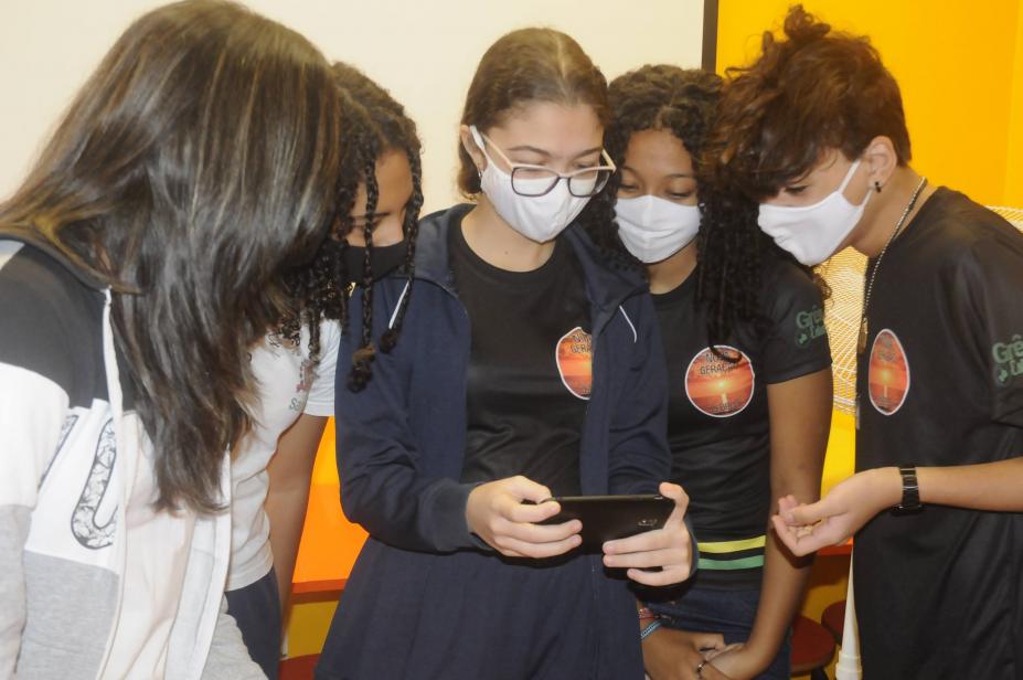 #pracegover Foto mostra quatro alunas e um aluno observando vídeo em celular na mão de uma das alunas
