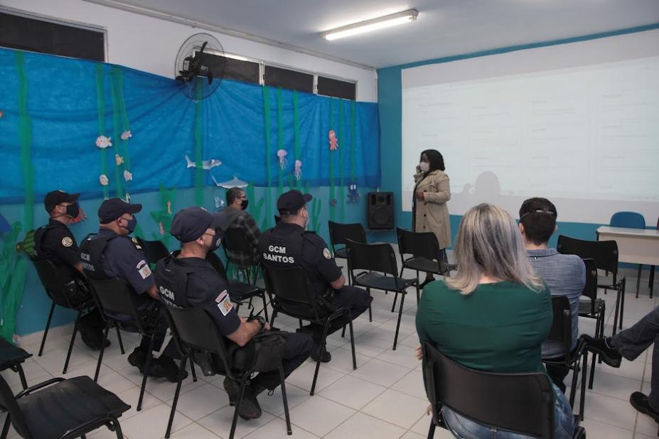 sala de aula com guardas municipais uniformizados sentados, e outras pessoas. Ao fundo, uma mulher está em pé falando ao grupo. #paratodosverem