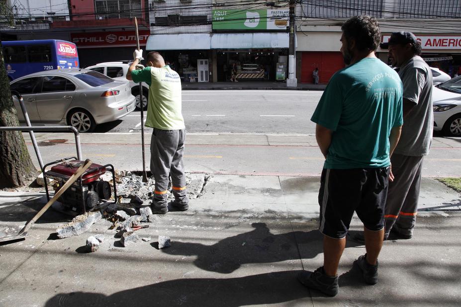 Homem está cavando buraco em trecho da calçada para instalação de barra. #Pracegover