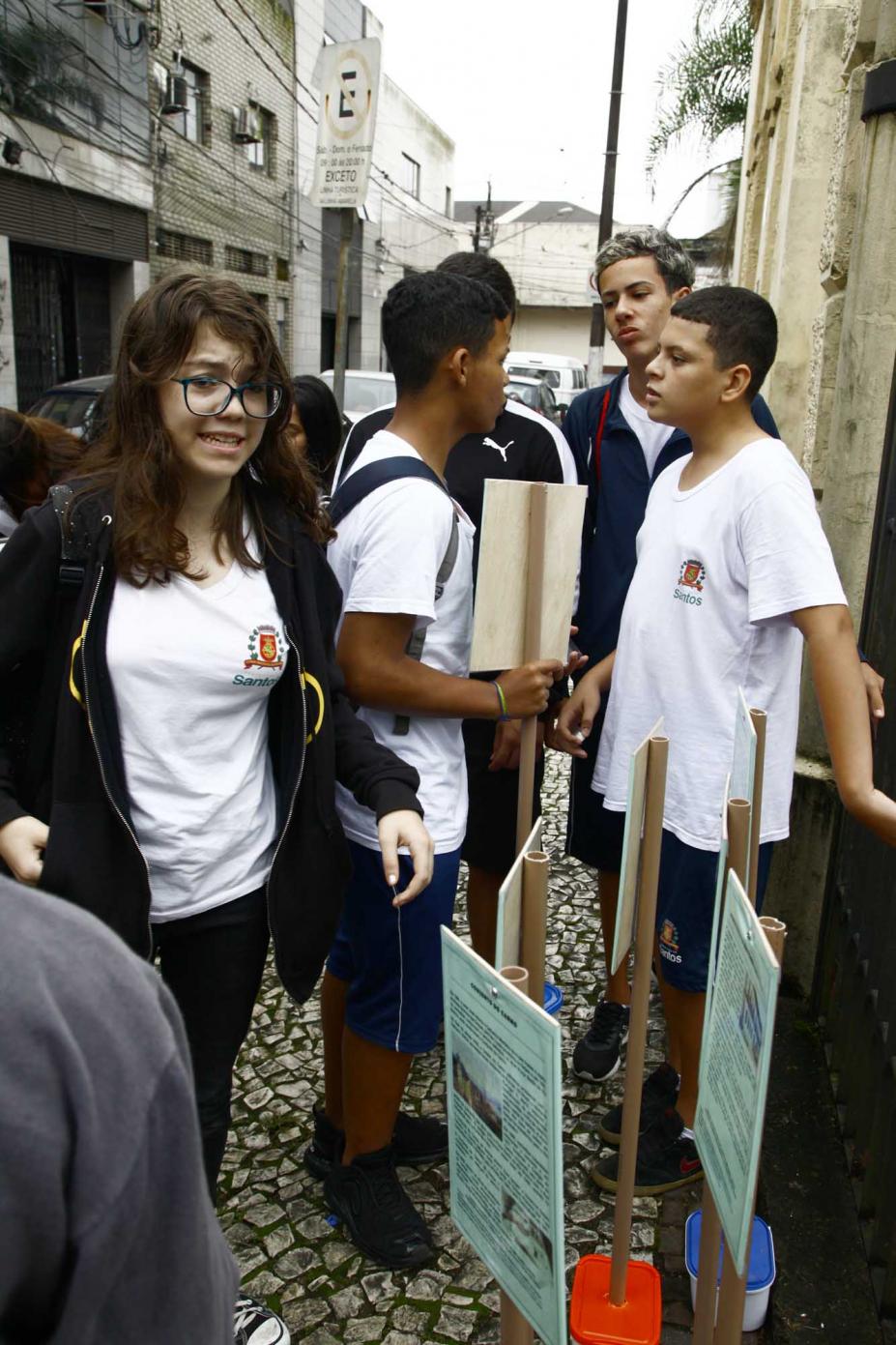 Alunos uniformizados estão em rua diante de placas informativas. #Pracegover