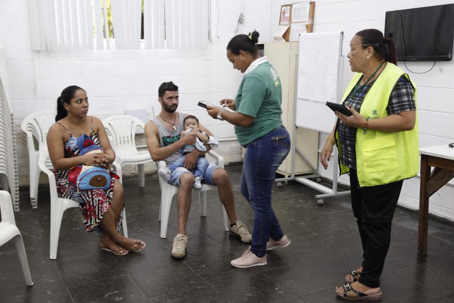 Agentes da ouvidoria estão em pé em salão conversando com um casal que está sentado. O homem está segurando um bebê no colo. #Paratodosverem