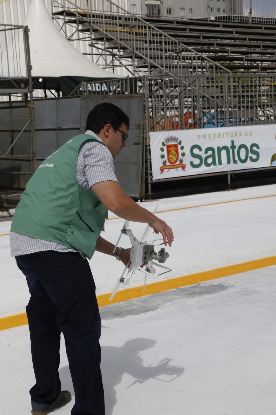 Homem vestindo colete de agente de combate à dengue  segura drone no meio do sambódromo.  #Pracegover