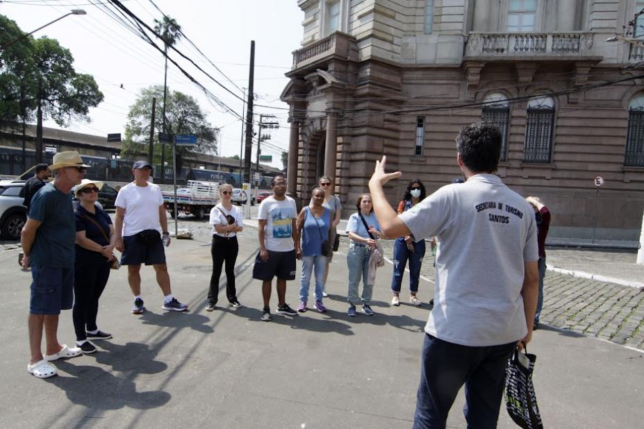 guia usando camiseta da seectur está falando a grupo de pessoas em frente à Bolsa Oficial do Café. #paratodosverem 