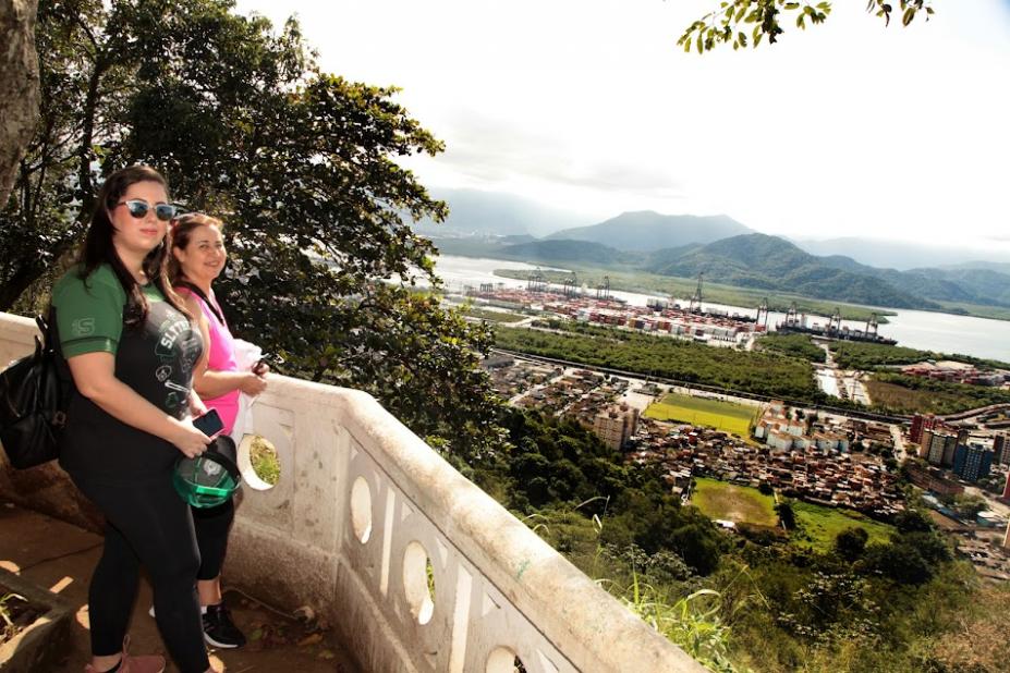 duas pessoas estão em pé atrás de mureta de santos no alto de um morro. Abaixo, vista ampla da cidade. #paratodosverem