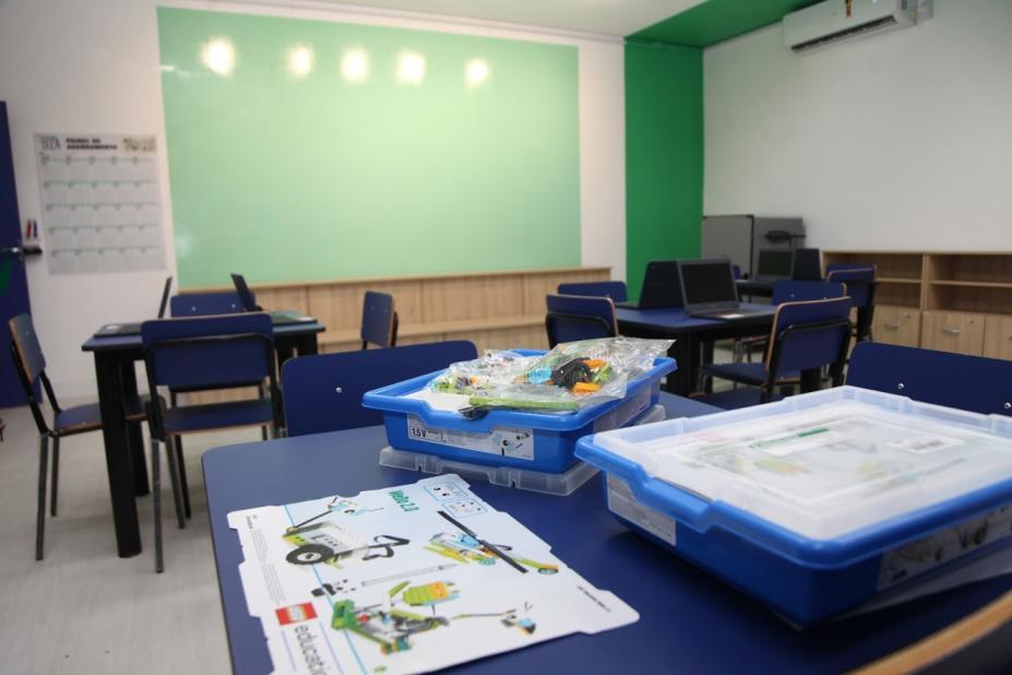 Sala de aula com mesa em primeiro plano. Sobre ela há duas caixas e um desenho. Ao fundo, a lousa. #Paratodosverem