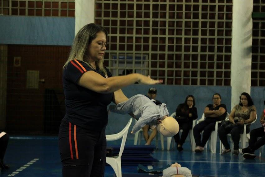 mulher uniformizada segura boneco de bebê e simula movimento para desengasgamento. Público está ao fundo. #paratodosverem