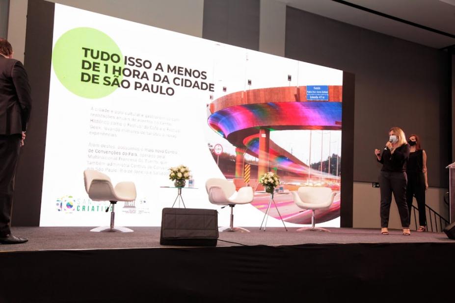 secretária fala no palco com viaduto de santos aparecendo no painel #paratodosverem