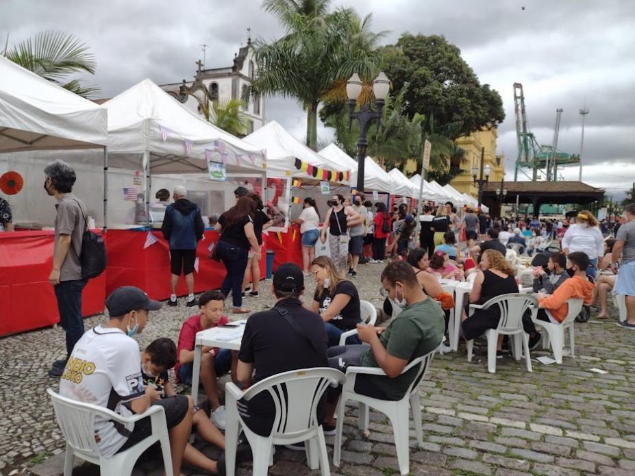 Barracas ao fundo e pessoas sentadas em mesa na rua #paratodosverem