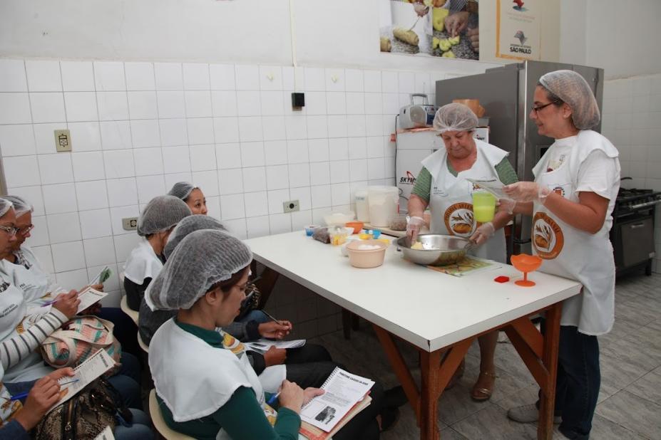 Duas cozinheiras preparam pão enquanto alunas observam sentadas #paratodosverem