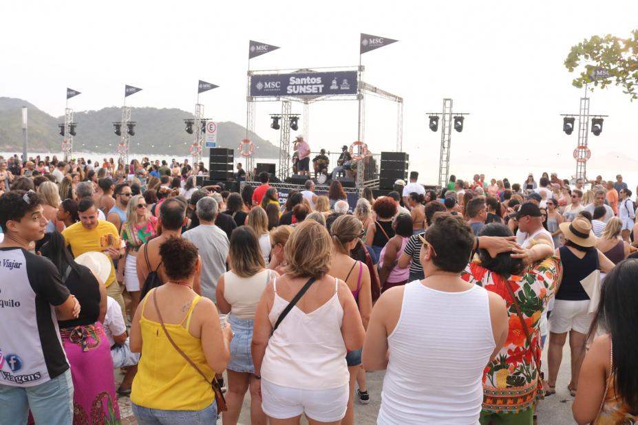 pessoas assistem show na ponta da praia com navio ao fundo. #paratodosverem