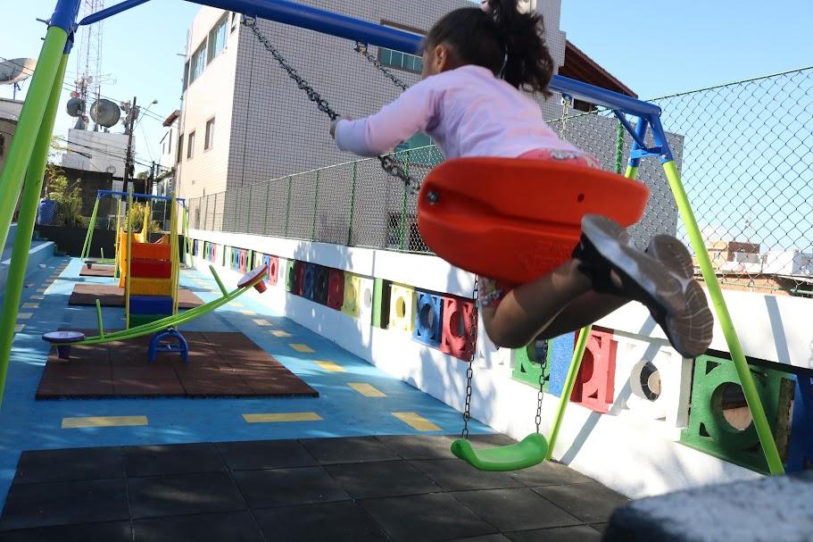 menina está brincando em balancê. Seus pés e corpo estão no ar, em primeiro plano e no alto. Ao fundo, os novos brinquedos. #paratodosverem