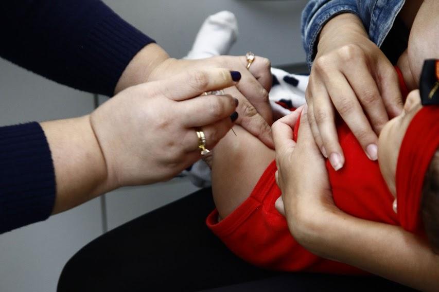 criança está no colo sendo vacinada pelas  mãos de uma  mulher. #pratodosverem