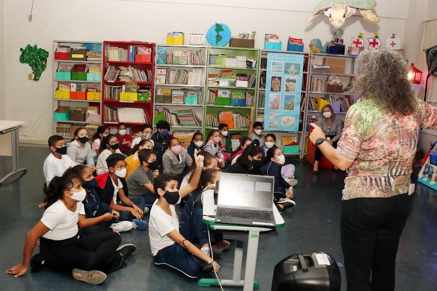 ALUNOS ESTÃO SENTADOS NO CHÃO ATENTOS A UMA MULHER QUE FALA EM PÉ. uM DELES LEVANTA A MÃO PARA PARTICIPAR. A sala tem prateleiras com livros e decoração voltada para o meio ambiente. #paratodosverem