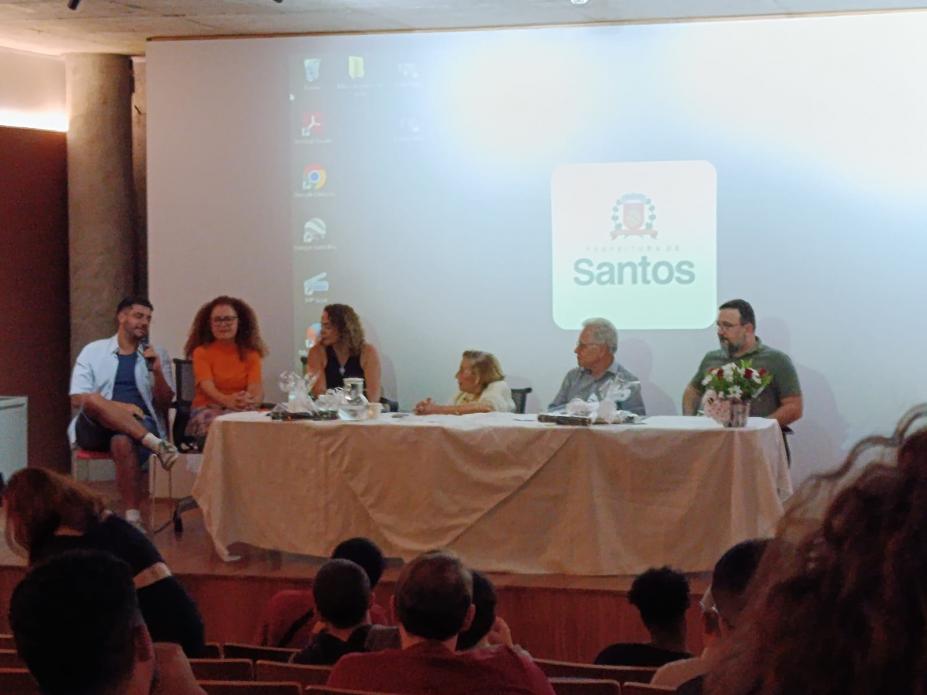 mesa de debates com seis pessoas sentadas e Wilma ao centro. #paratodosverem