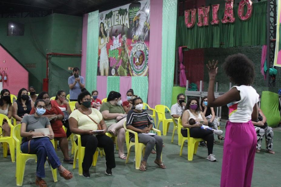 Mulheres sentadas observam instrutora em palestra na quadra da escola de samba União Imperial. #pratodosverem