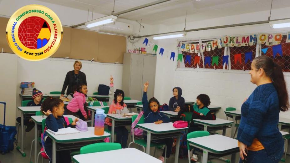 sala de aula com alunos e professores à frente. Selo está sobreposto sobre a imagem, no alto, à esquerda. #paratodosverem 