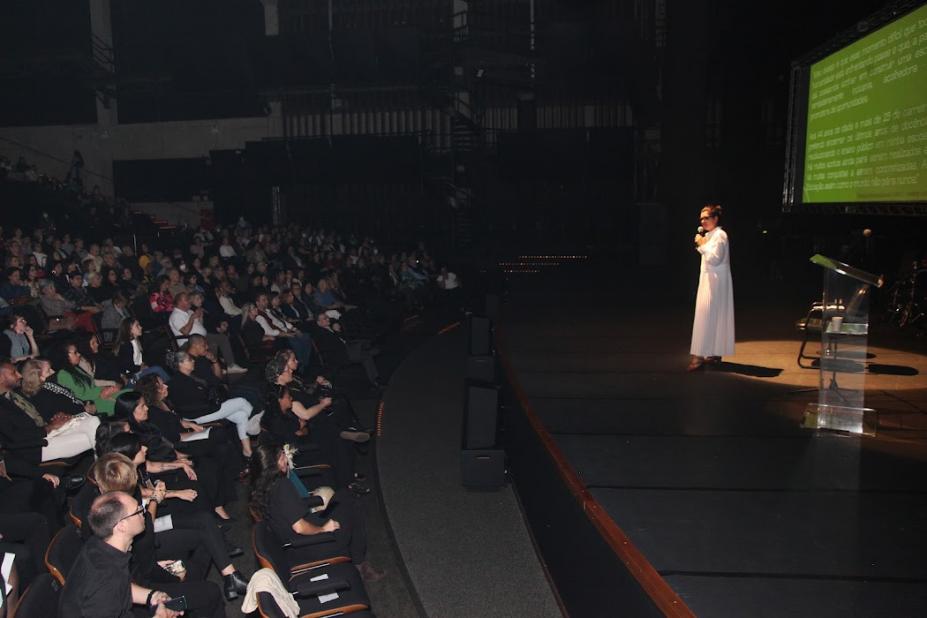 Especialista, no palco do teatro, fala ao microfone para o público na plateia formado por educadores. #paratodosverem
