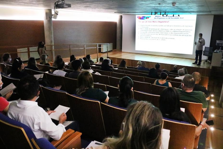 auditório com pessoas sentadas acompanhando palestra. Ao fundo há um telão. #paratodosverem