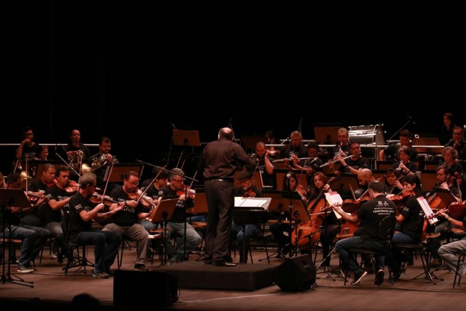 músicos estão sentados tocando seus instrumentos. regente está à frente do grupo, de costas para a foto. O palco está escuro. #paratodosverem