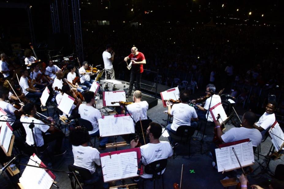 Músicos da Sinfônica de Santos estão em palco tocando diante de suas partituras em estantes. O maestro está ao fundo. Mais ao fundo, no escuro, se vê algumas luzes. #Pracegover