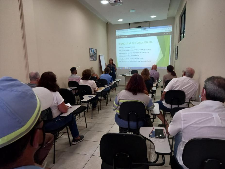 palestrante na frente da sala com telão ao fundo e pessoas assistindo #paratodosverem