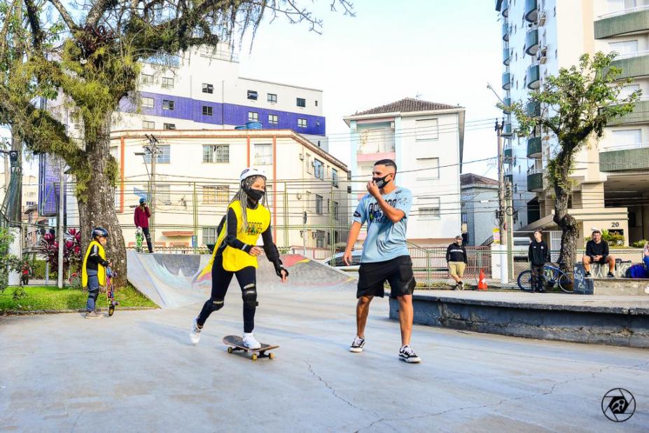 instrutor e menina uniformizada sobre skate estão na pista. Há árvores e prédios ao fundo. #paratodosverem