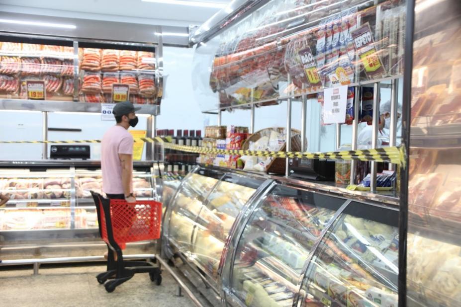 Homem está diante de balcão de carnes em supermercado. Ele está ao lado de um carrinho de mercadorias. O balcão está demarcado por faixa zebrada. #paratodosverem