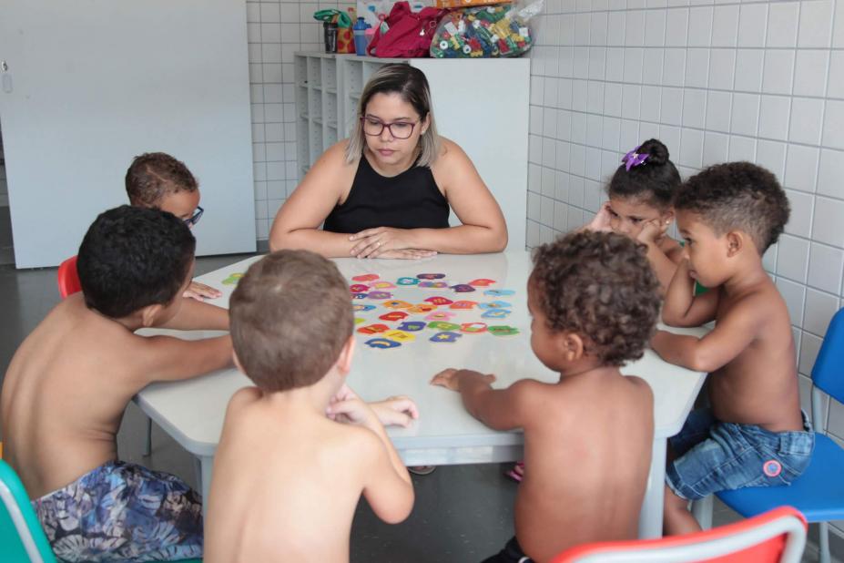 Crianças estão sentadas em torno de uma mesa. Elas estão jogando um jogo acompanhadas por uma mulher. #Pracegover