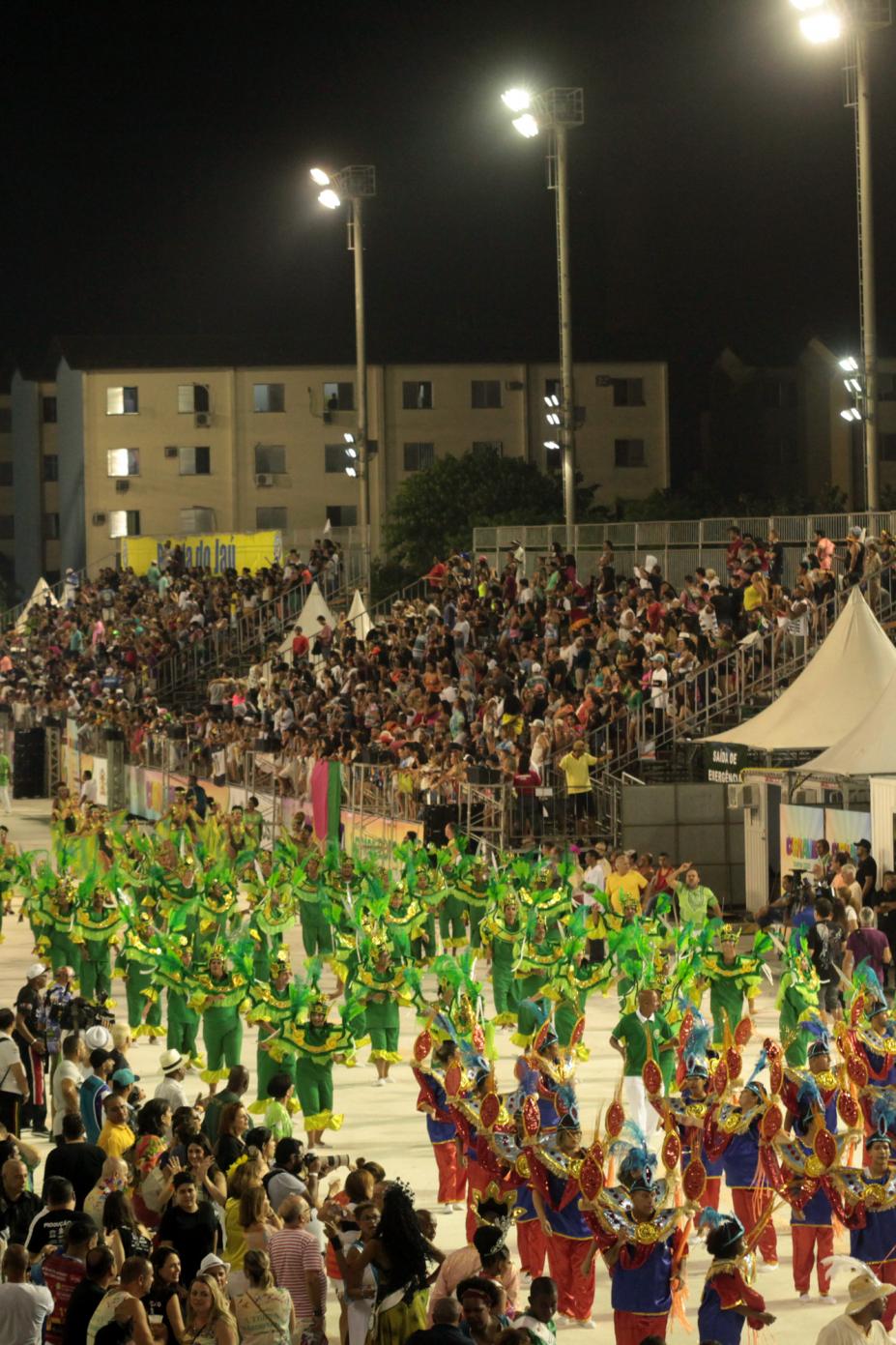 Desfile de escola de samba em passarela, com o público na arquibancada ao fundo. #Paratodosverem