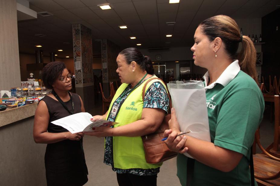 Agentes da Ouvidoria vestidas com colete e camiseta de identificação conversam com uma mulher em um estabelecimento comercial. #Pracegover