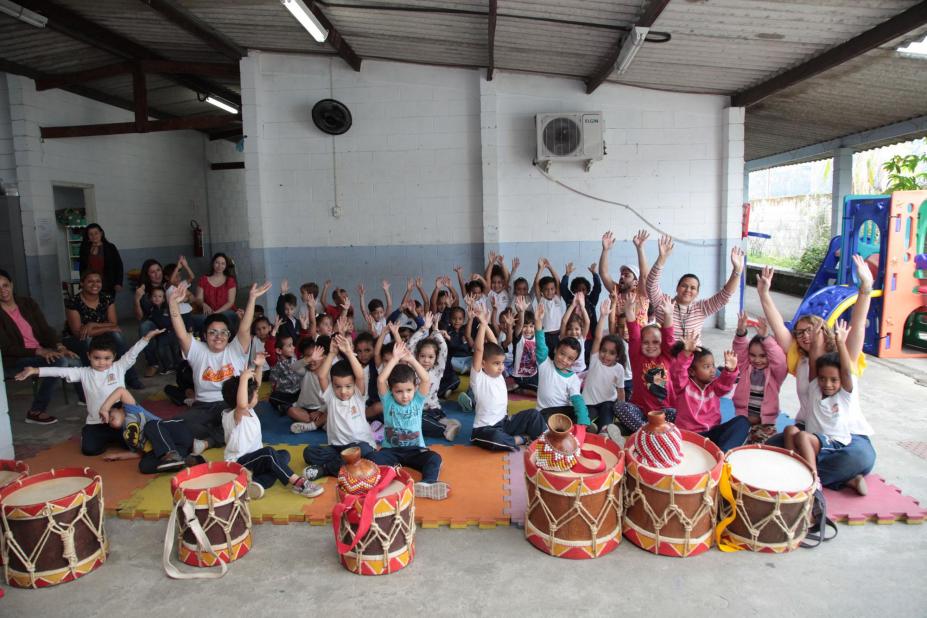 Crianças estão em pátio escolas, todas com as mãos para o alto. À frente do grupo há diversos tambores musicais. #Pracegover