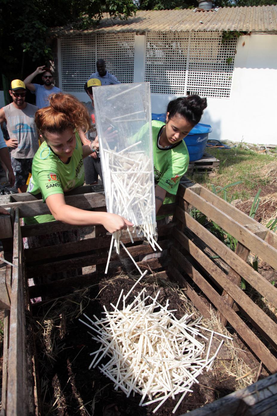 Duas mulheres seguram recipiente e depositam o conteúdo em uma composteira. #Paratodosverem