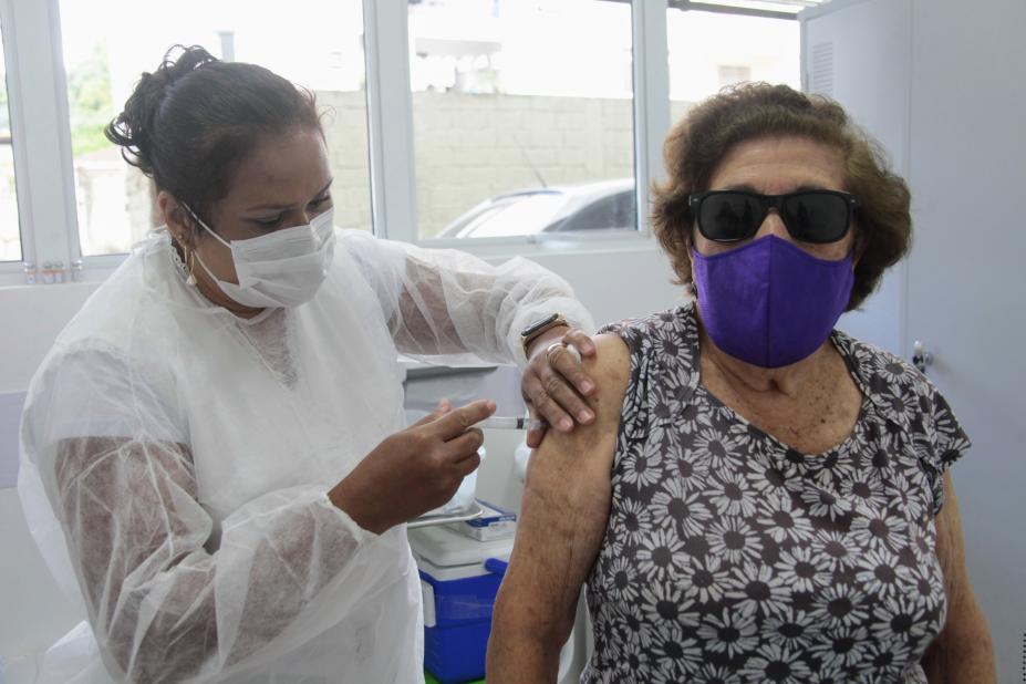 Enfermeira usando máscara aplica vacina em braço direito de uma mulher que também usa máscara #paratodosverem