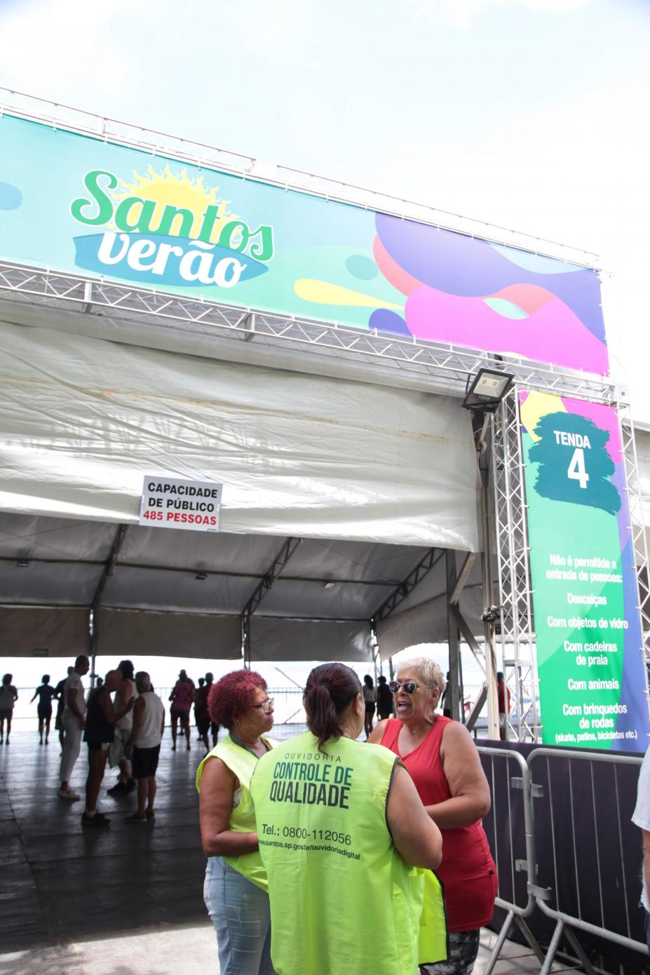 Agentes da Ouvidoria conversam com uma mulher na entrada de tenda de verão. #Pracegover