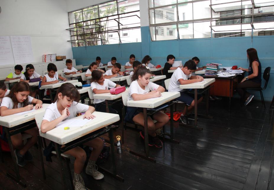 Alunos estão em sala de aula sentados e fazendo prova. Professora está no canto direito da imagem, com sua mesa próxima à parede da sala. #Pracegover