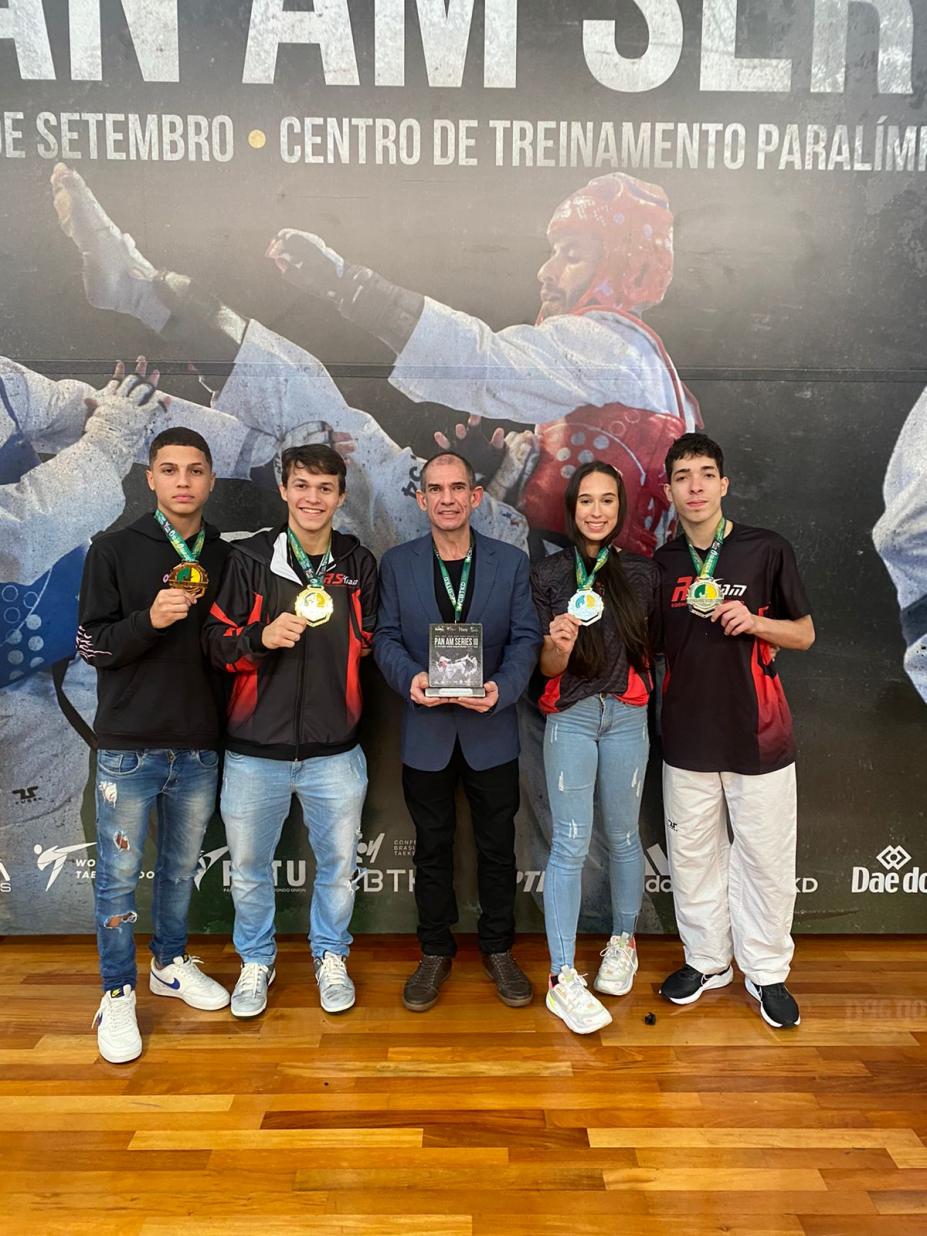 Treinador e atletas exibem medalhas e troféu em frente a banner do evento. #paratodosverem