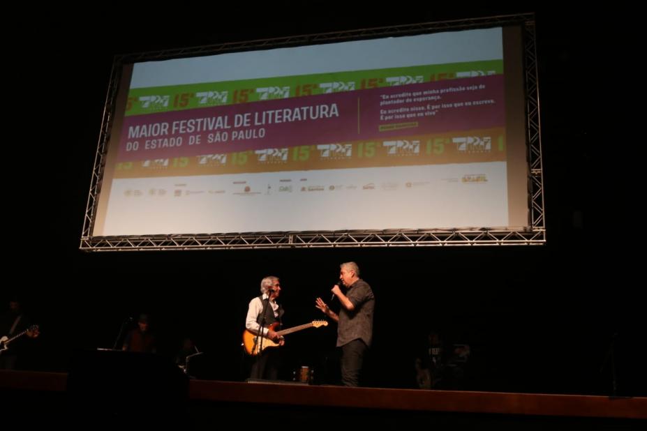 cantor segura guitarra no palco ao lado de um homem. Ao fundo e no alto, um telão onde se lê Tarrafa Literária e a programação. #paratodosverem 