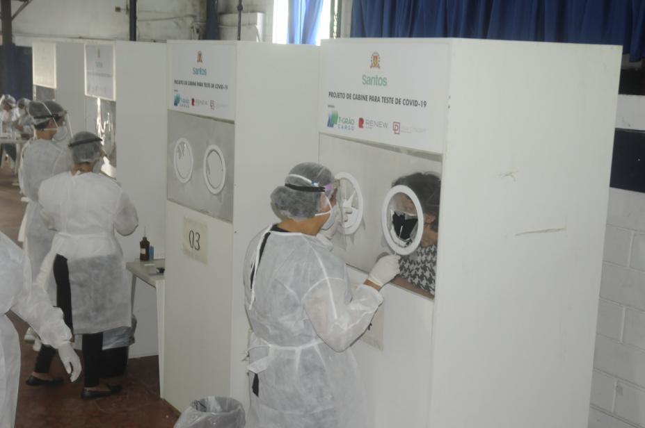 pessoas estão passando por teste nasal de covid atrás de biombos. Técnicos vestidos de branco procedem o exame do lado externo. #paratodosverem