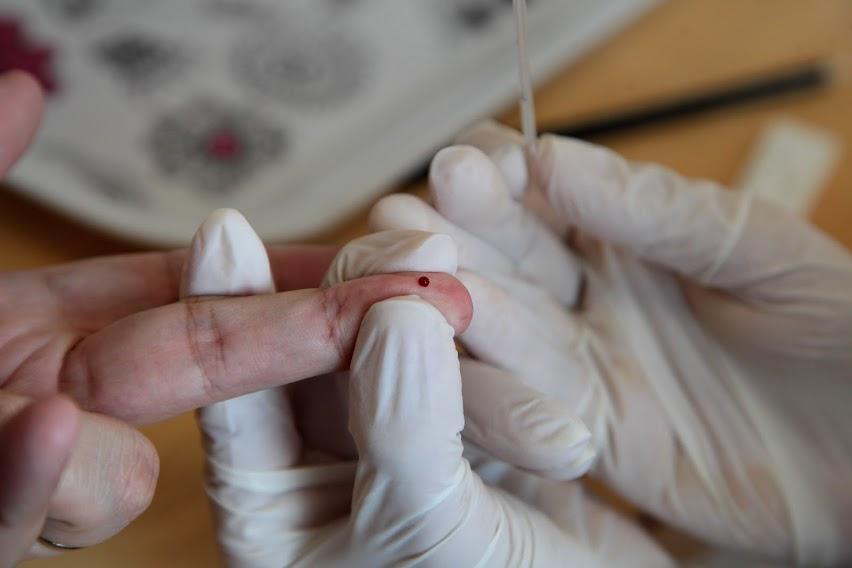 mãos com luvas seguram o dedo de uma pessoa que tem uma gota de sangue. #paratodosverem