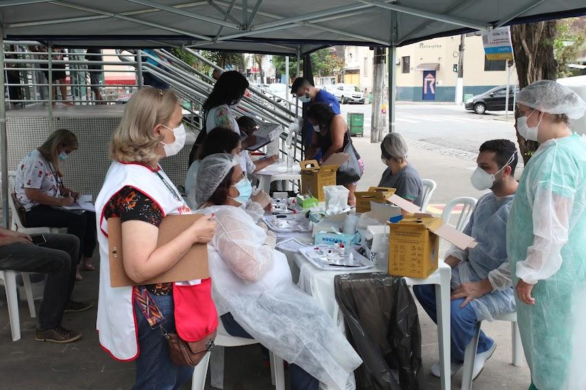 movimentação sob tenda, com pessoas em pé e sentadas usando avental, touca e máscara. #paratodosverem