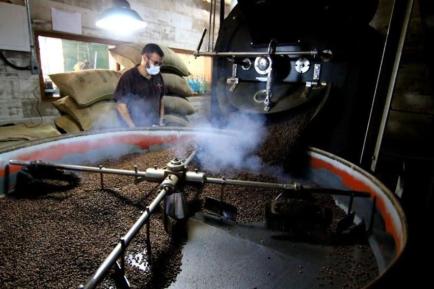 Equipamento de torra de café com diversos grãos. Máquina está em movimento. Homem está ao fundo diante de sacas de café empilhadas. #paratodosverem