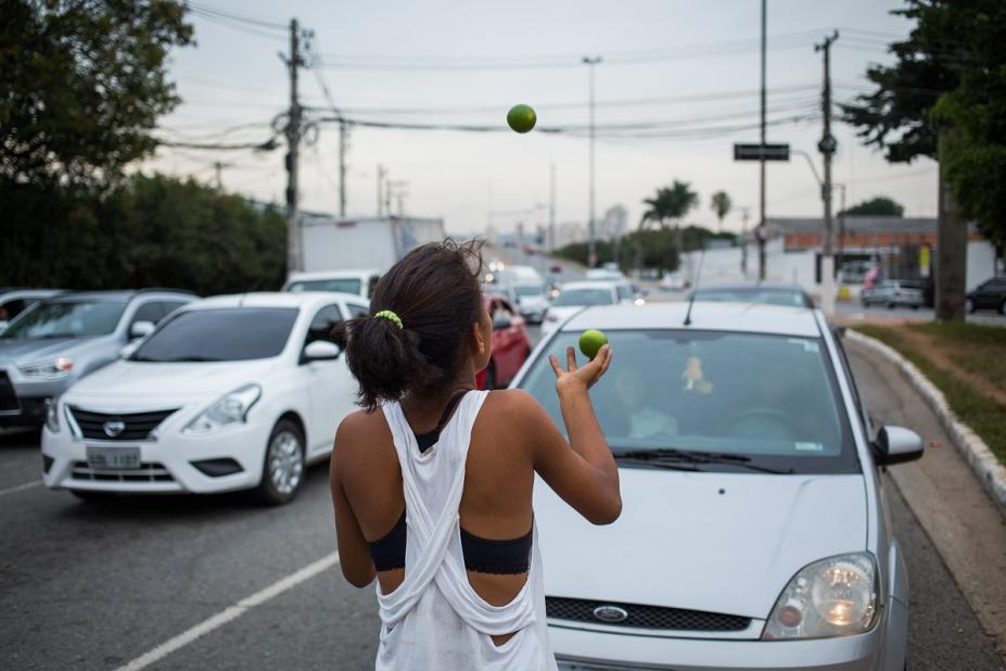 menina de costas para a foto faz malabares com limões na avenida diante de carros. #paratodosverem