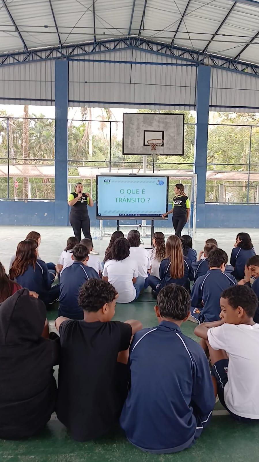 alunos sentados em quadra assistem apresentação sobre trânsito. #paratodosverem