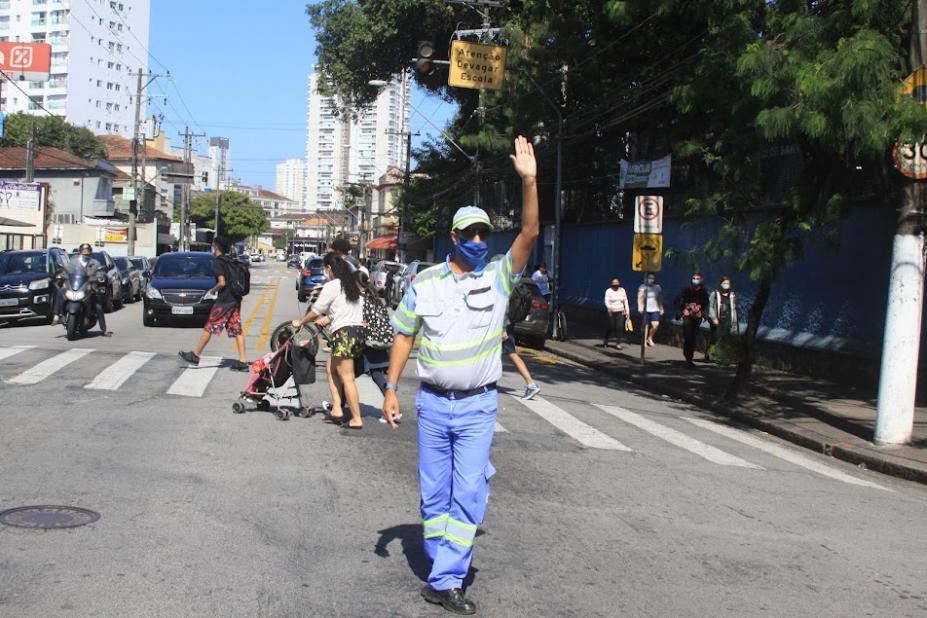 agente da CET está na rua, à frente de uma faixa de pedestres. Ele está com o braço esquerdo para o alto, fazendo gesto para veículos de 'pare'. Ao fundo, pessoas atravessam a rua. #paratodosverem