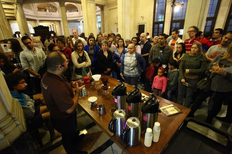 Várias pessoas assistem à preparação de café. Homem que está demonstrando fica atrás de mesa onde estão várias garrafas térmicas, coadores e copinhos de café. #paratodosverem