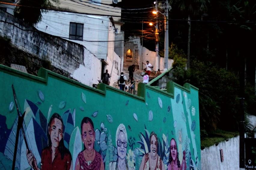 MURO ILUSTRADO NO SOPÉ DO MONTE SERRTA COM A ESCADARIA NO ALTO E PESSOAS CIRCULANDO. #PARATODOSVEREM