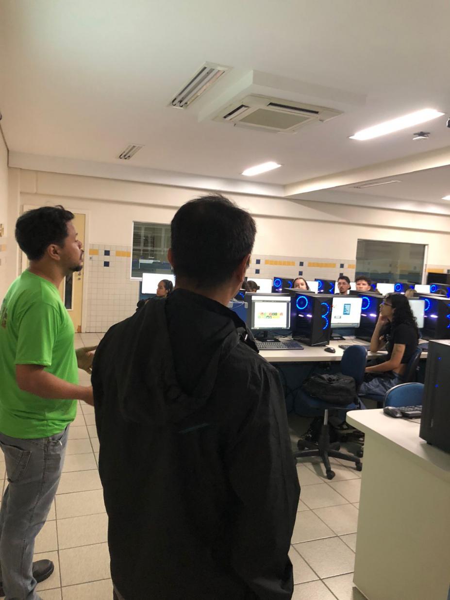 duas pessoas em pé e alunos sentados na frente de computadores #paratodosverem